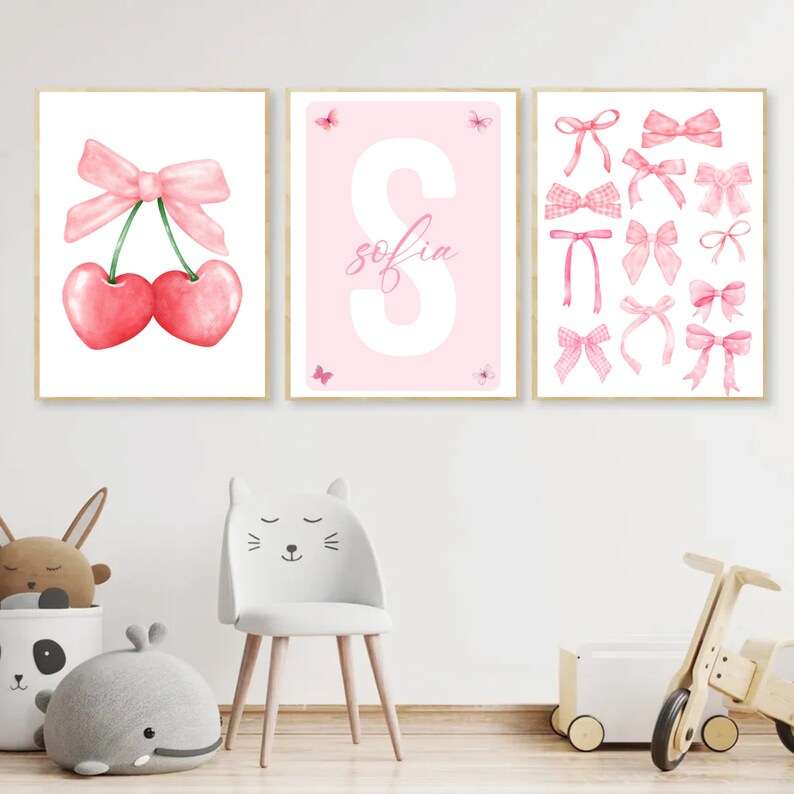May include: Three pink and white watercolor prints for a nursery or child's room. The first print features two red cherries with a pink bow. The second print features a large white letter S with the name "sofia" written in cursive. The third print features a collection of pink bows in various styles.