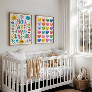May include: Two framed art prints with gold frames hang above a white crib. One print says "YOU ARE MY SUNSHINE" with colorful flowers and a sun. The other print features rows of colorful hearts. A teddy bear and a blanket are in the crib.