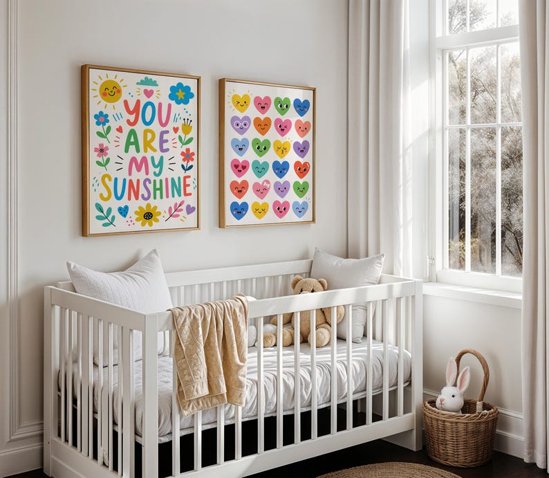May include: Two framed art prints with gold frames hang above a white crib. One print says "YOU ARE MY SUNSHINE" with colorful flowers and a sun. The other print features rows of colorful hearts. A teddy bear and a blanket are in the crib.