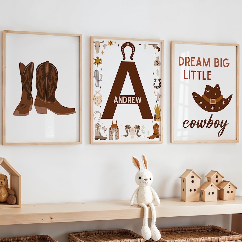 May include: Three framed prints with a western theme. The first print features a pair of brown cowboy boots. The second print features the letter A with the name Andrew below it and is surrounded by western themed icons. The third print features a brown cowboy hat with the text "Dream Big Little Cowboy".