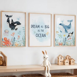 May include: Three framed watercolour prints with a marine theme. One features a black and white orca, another reads "Dream as Big as the Ocean," and the third depicts a blue whale. The prints are in light wood frames, suitable for a nursery or child's room.