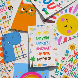 May include: A collection of colorful, illustrated cards with positive affirmations and cheerful designs. The cards feature images of a cat, dinosaur, sun, and rainbow, with phrases like "I am loved" and "You are my sunshine."