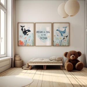 May include: Three framed watercolour prints with ocean themes. One features a black and white orca, another reads "Dream as big as the ocean," and the third depicts a blue whale. The prints are displayed above a small wooden daybed and a brown teddy bear.