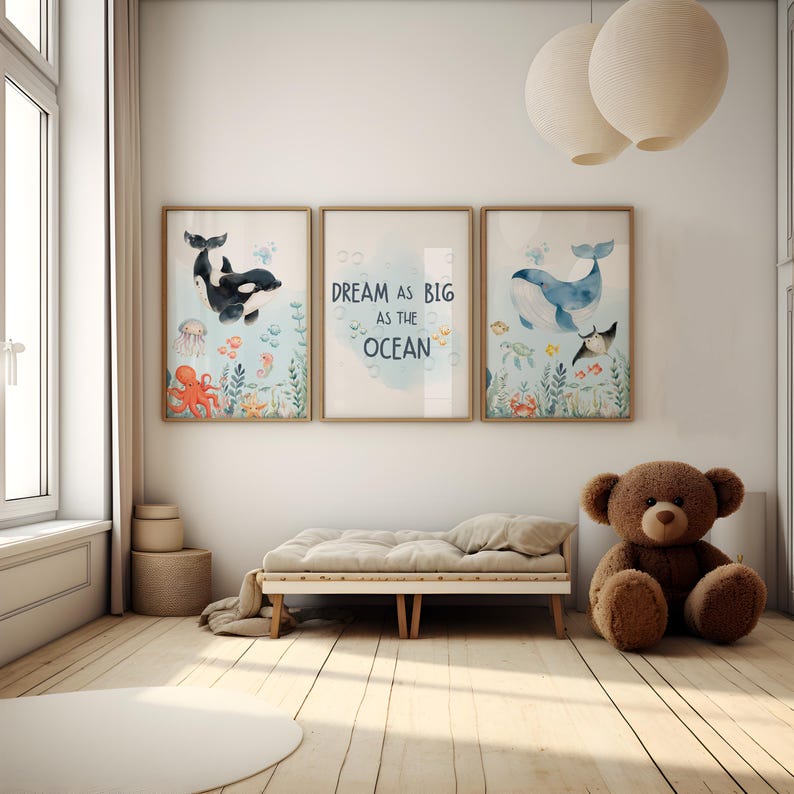May include: Three framed watercolour prints with ocean themes. One features a black and white orca, another reads "Dream as big as the ocean," and the third depicts a blue whale. The prints are displayed above a small wooden daybed and a brown teddy bear.