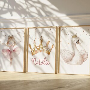 May include: Three framed watercolor prints featuring a ballerina, a crown with the name "Natalie", and a swan wearing a crown. The prints have a soft, pastel color palette with gold star accents, and are set in wooden frames.