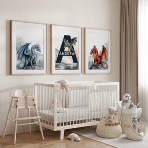 May include: Three framed art prints featuring watercolor dragons in blue, green, and red. One print has the name "Andrew" with a large letter "A". The prints are displayed above a white crib in a nursery setting.