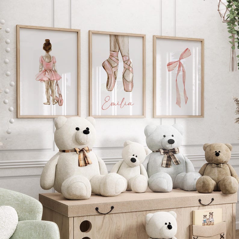 May include: Three framed watercolor prints with a ballerina theme. The prints feature a ballerina in a pink tutu, a pair of pink ballet shoes, and a pink ribbon. The prints are hung on a white wall above a shelf with stuffed teddy bears.