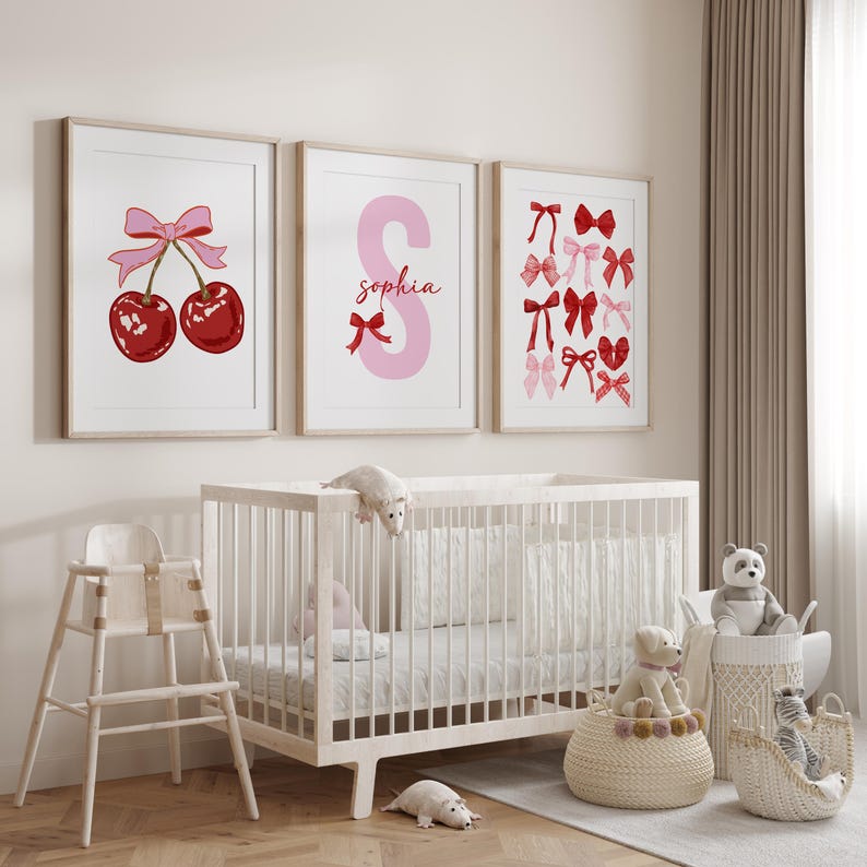 May include: Three framed prints on a white wall above a white crib. The prints feature a red cherry with a pink bow, a pink letter S with the name "Sophia" and a collection of red and pink bows.