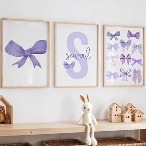May include: Three framed prints with a purple watercolour bow theme. The first print features a large purple bow. The second print features a large purple letter S with the name "sarah" written below. The third print features a collection of smaller purple bows.
