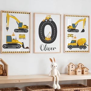 May include: Three framed prints with a construction theme. The prints display yellow construction vehicles, such as excavators and a cement mixer, on a white background. One print has the name "Oliver" in cursive. Each print is framed in a light wood frame.