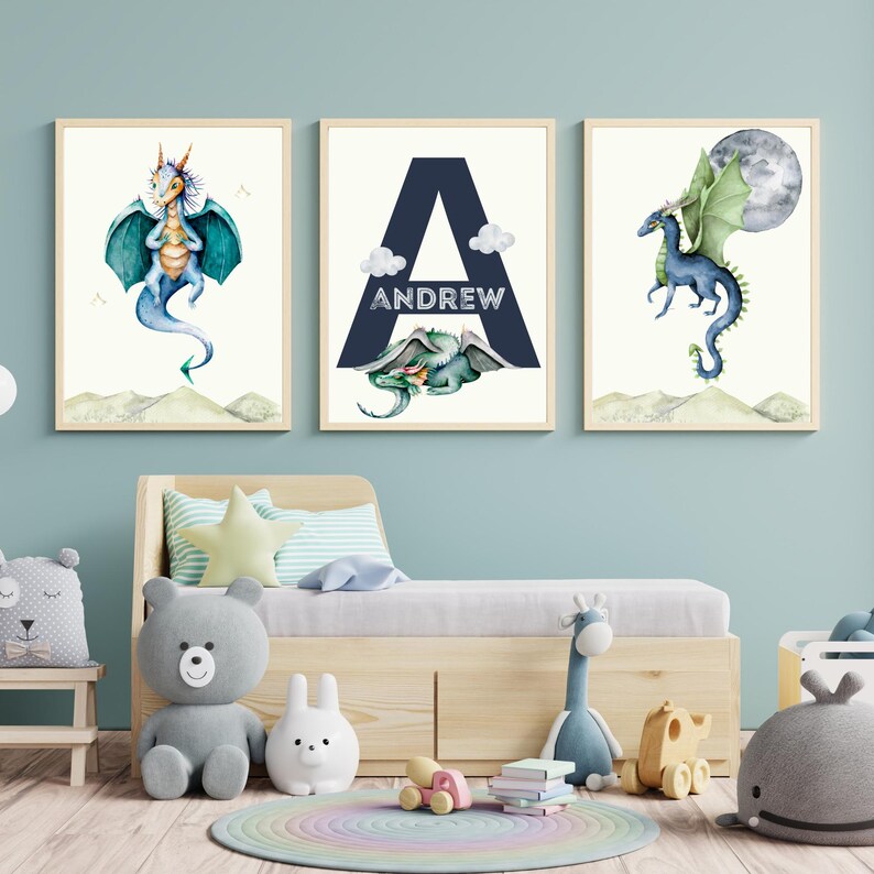 May include: Three watercolor prints featuring a blue dragon, the letter A with the name Andrew, and a blue dragon with a green wing. The prints are framed and hung on a light blue wall above a wooden bed.