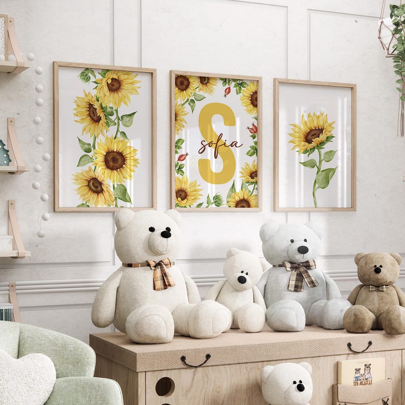 May include: Three framed prints with watercolor illustrations of sunflowers. The middle print features a large yellow letter "S" with the name "sofia" written in brown script. The prints are displayed on a white wall above a wooden shelf with stuffed teddy bears.