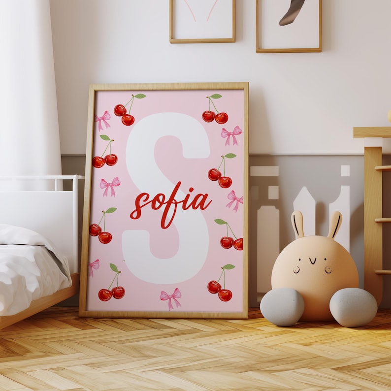 K&ouml;nnte beinhalten: Ein gerahmter rosa Druck mit dem Namen "Sofia" in roter Schrift und einem gro&szlig;en wei&szlig;en "S". Der Druck ist mit roten Kirschen und rosa Schleifen verziert. Der Rahmen ist aus hellem Holz. Der Druck wird in einem Kinderzimmer ausgestellt.