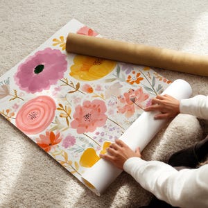 May include: Floral wallpaper roll with a watercolor design featuring pink, yellow, and orange flowers. A brown cardboard tube rests on top. The wallpaper is unrolled on a light-colored carpet.