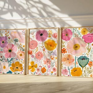 May include: Three framed floral art prints featuring colorful watercolor flowers in pink, yellow, orange, and purple. The botanical illustrations are set against a white background, with wooden frames. The artwork is leaning against a white wall.