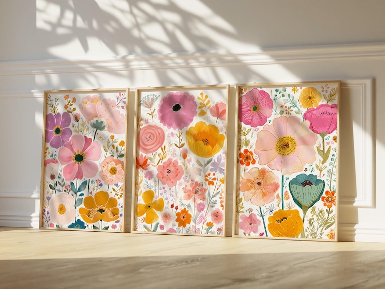 May include: Three framed floral art prints featuring colorful watercolor flowers in pink, yellow, orange, and purple. The botanical illustrations are set against a white background, with wooden frames. The artwork is leaning against a white wall.