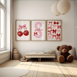 May include: Three framed prints with a pink and red color scheme. The first print features a pair of red cherries with a pink bow. The second print features a pink letter S with the name "Sophia" written below. The third print features a collection of pink and red bows.