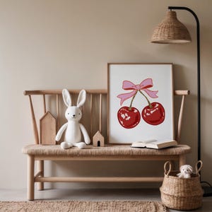 May include: A white bunny plush toy sits on a wooden bench with a framed print of two red cherries with a pink bow. The print is on a white background.