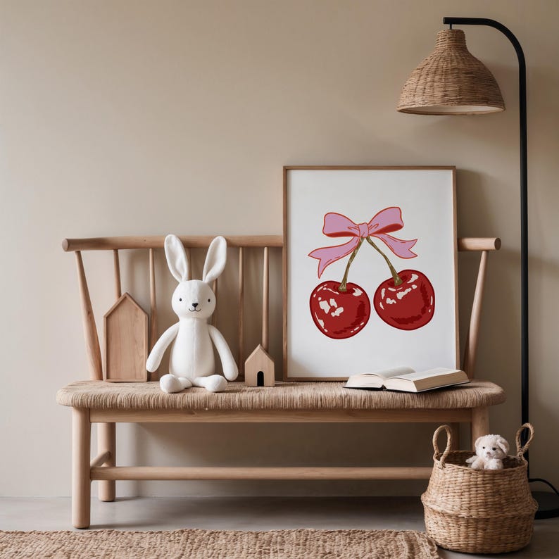 May include: A white bunny plush toy sits on a wooden bench with a framed print of two red cherries with a pink bow. The print is on a white background.