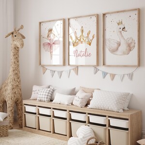 May include: Three framed watercolor prints with a ballerina, crown, and swan, in a child's room. The crown print has the name "Natalie" in cursive. A woven giraffe and storage unit with pillows and bins complete the scene.