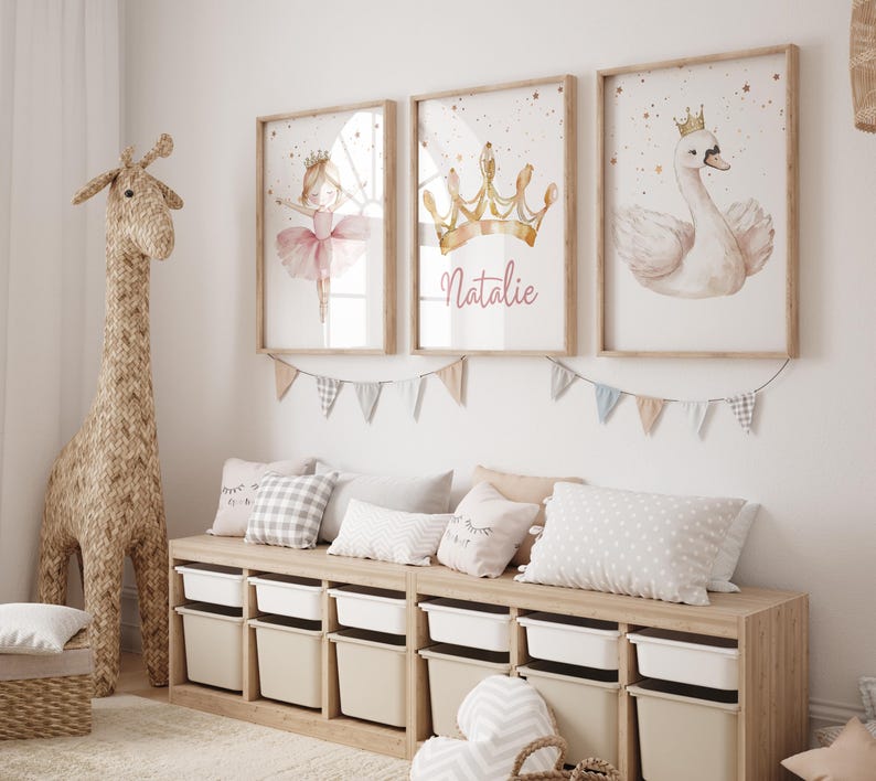 May include: Three framed watercolor prints with a ballerina, crown, and swan, in a child's room. The crown print has the name "Natalie" in cursive. A woven giraffe and storage unit with pillows and bins complete the scene.