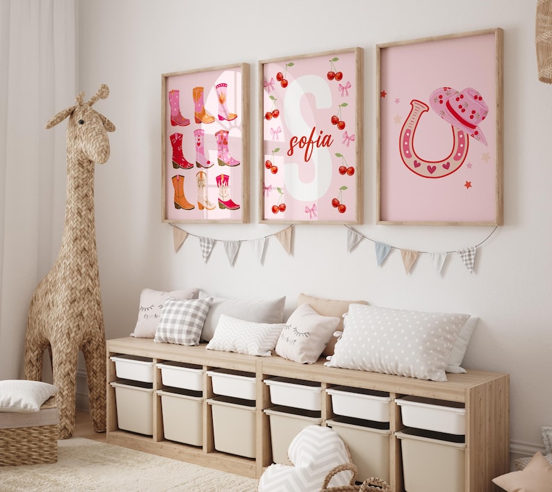 K&ouml;nnte beinhalten: Drei gerahmte rosa Wandkunstdrucke. Einer zeigt bunte Cowboy-Stiefel, ein anderer hat den Namen "Sofia" mit Kirschen und der dritte zeigt ein Hufeisen und einen Cowboyhut. Die Drucke befinden sich &uuml;ber einem Holzaufbewahrungsschrank mit wei&szlig;en Beh&auml;ltern.
