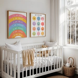 May include: A white crib with a beige blanket and a teddy bear sits in a bright room. Two framed prints hang above the crib. One features a rainbow with positive affirmations, and the other displays various facial expressions.