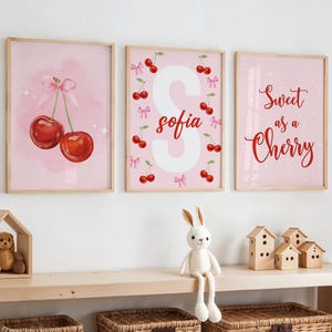 Puede incluir: Tres láminas enmarcadas con temática de cerezas rosas. Una lámina presenta dos cerezas con un lazo rosa. Otra tiene el nombre "Sofia" con cerezas y lazos. La tercera lámina dice "Sweet as a Cherry".