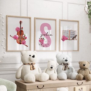 May include: Three framed prints with watercolor illustrations of a violin, the letter S with the name Sophia, and a book with a feather.  The prints are hanging on a white wall above a wooden shelf with four teddy bears.