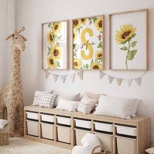 May include: Three framed prints with watercolor sunflower designs. The center print features a large yellow letter "S" with the name "Sofia" written below it. The prints are hung on a white wall above a wooden bench with storage bins.