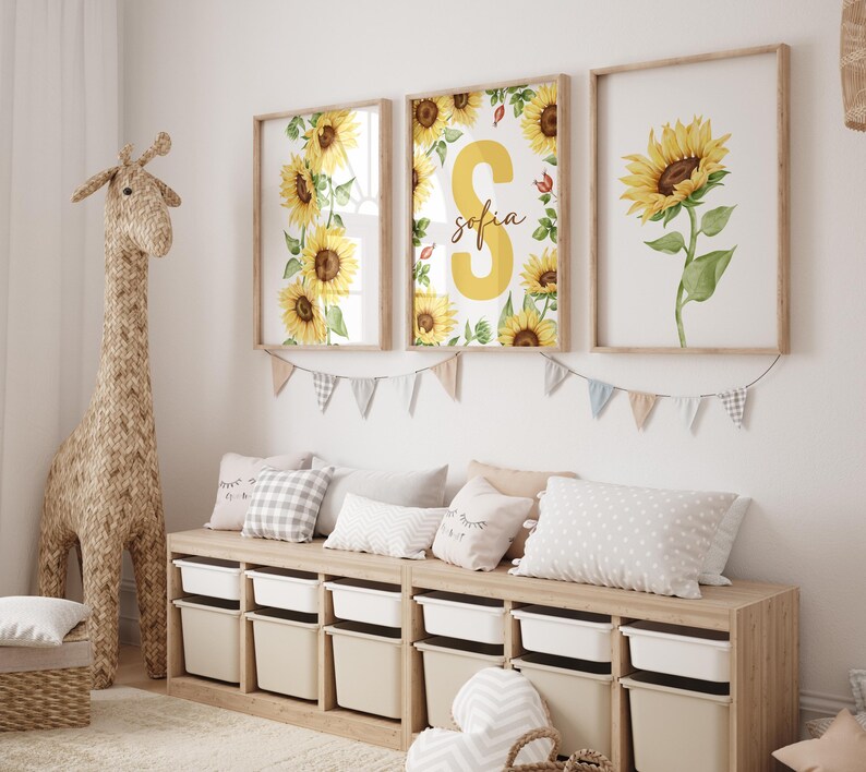 May include: Three framed prints with watercolor sunflower designs. The center print features a large yellow letter "S" with the name "Sofia" written below it. The prints are hung on a white wall above a wooden bench with storage bins.