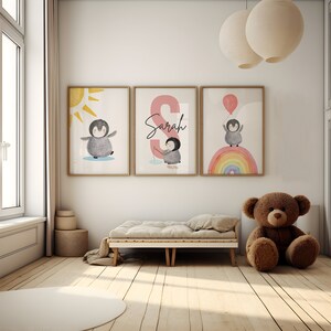 May include: Three framed prints with a penguin theme. The first print features a penguin standing on a white background with a yellow sun behind it. The second print features a pink letter S with the name Sarah written in black. The third print features a penguin standing on a rainbow with a blue balloon in its hand.
