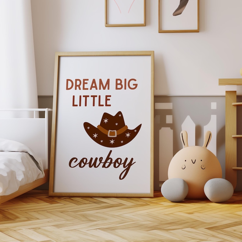 May include: A white poster with brown text that reads "Dream Big Little Cowboy" with a brown cowboy hat with white stars and a brown belt illustration.