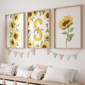 May include: Three framed prints with a sunflower theme. The center print features a large yellow letter "S" with the name "sofia" written below it. The prints are hung on a white wall with a string of flags.