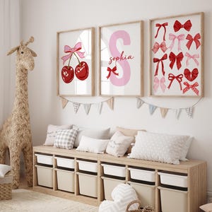 May include: Three framed prints for a child's room. The first print features a pink bow and two red cherries. The second print features a pink letter S with the name "Sophia" written below. The third print features a variety of red and pink bows.