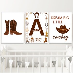 May include: Three brown and white nursery prints with a cowboy theme. The first print features a pair of brown cowboy boots. The second print features the letter A with the name Andrew written inside. The third print features a brown cowboy hat with the text "Dream Big Little Cowboy".
