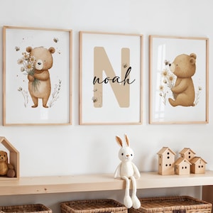 Może przedstawiać: Trzy oprawione wydruki artystyczne przedstawiające akwarelowego misia trzymającego kwiaty, dużą literę "N" z imieniem "Noah" i misia z kwiatami. Wydruki mają jasne drewniane ramy i są ozdobione pszczołami. Pluszowy królik i drewniane domki znajdują się na półce poniżej.