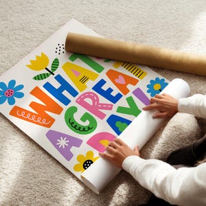 May include: A colorful poster with the words "WHAT A GREAT DAY" in large, playful letters. The poster features floral illustrations and is being unrolled. A brown cardboard tube rests nearby.