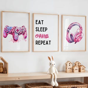 May include: Three framed prints with a gaming theme. One print shows a pink and blue video game controller. Another has the text "EAT SLEEP GAME REPEAT". The third print features pink headphones.