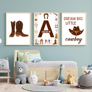 May include: Three printable wall art prints with a cowboy theme. The first print features a pair of brown cowboy boots. The second print features the letter A with the name Andrew and is surrounded by cowboy-themed illustrations. The third print features a brown cowboy hat and the text "Dream Big Little Cowboy".