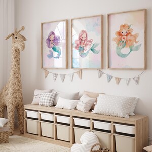May include: Three framed watercolor prints of mermaids with purple, pink, and orange hair. The prints are hung on a white wall above a wooden bench with pillows.