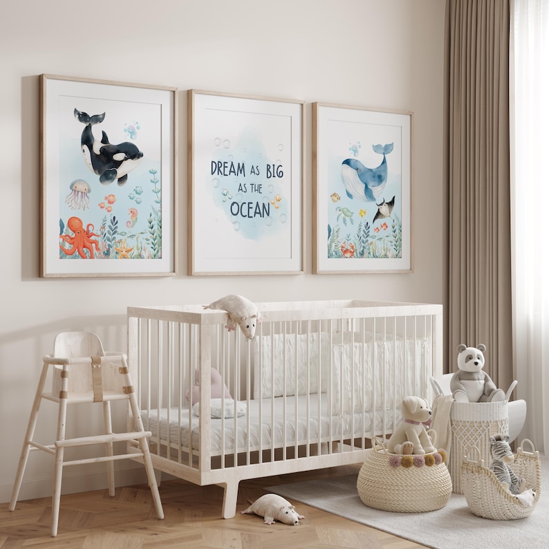 May include: Three framed watercolour prints with ocean themes hang above a white cot. The prints feature an orca, a whale, and the text "DREAM AS BIG AS THE OCEAN." A high chair, stuffed animals, and decorative baskets complete the nursery setting.