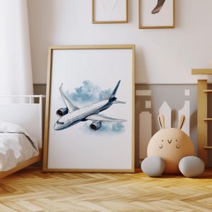Puede incluir: Un cuadro enmarcado con una ilustraci&oacute;n en acuarela de un avi&oacute;n de pasajeros volando por un cielo nublado. El avi&oacute;n es principalmente blanco con detalles azules. La obra est&aacute; en un marco de madera marr&oacute;n claro, para un dormitorio infantil.