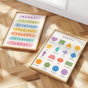 May include: Two framed educational prints on a wooden floor. One print displays the days of the week in colorful watercolor brushstrokes. The other print features various shapes with their names, including a heart, star, and diamond.