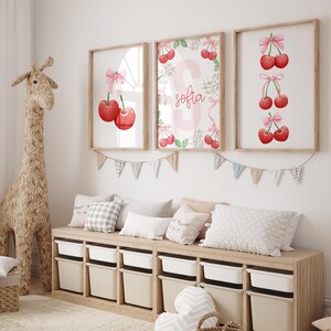 May include: Three framed prints with watercolor illustrations of cherries and the name "Sofia" in pink lettering. The prints are hung on a white wall above a wooden bench with cushions.