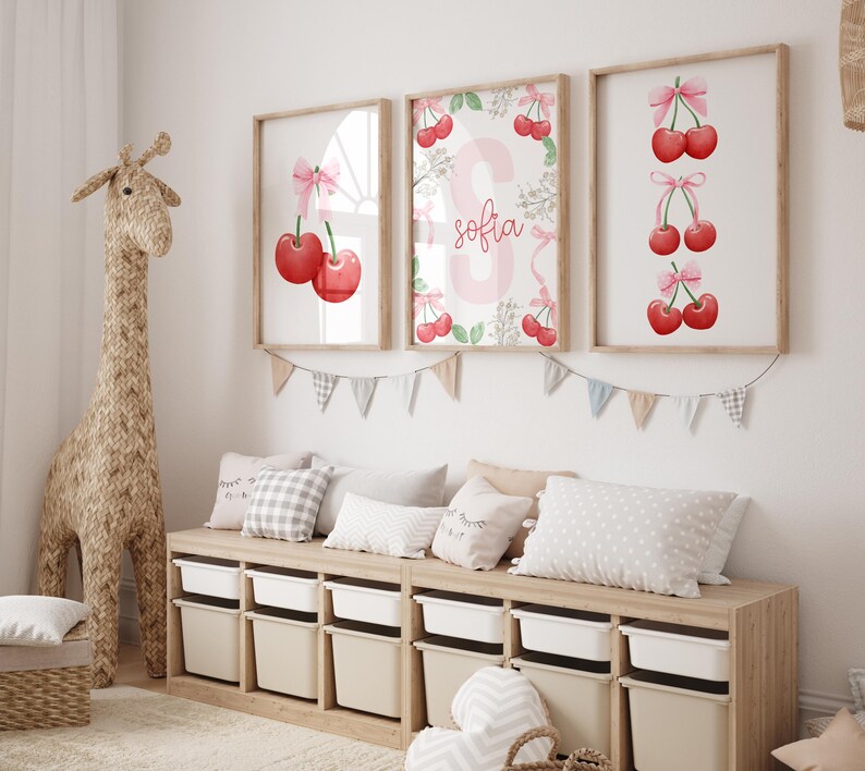 May include: Three framed prints with watercolor illustrations of cherries and the name "Sofia" in pink lettering. The prints are hung on a white wall above a wooden bench with cushions.
