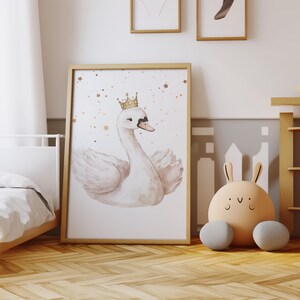 May include: A framed watercolor print of a swan wearing a gold crown, set against a backdrop of gold stars. The artwork is in a light wood frame. A plush bunny toy with gray feet sits on the floor.