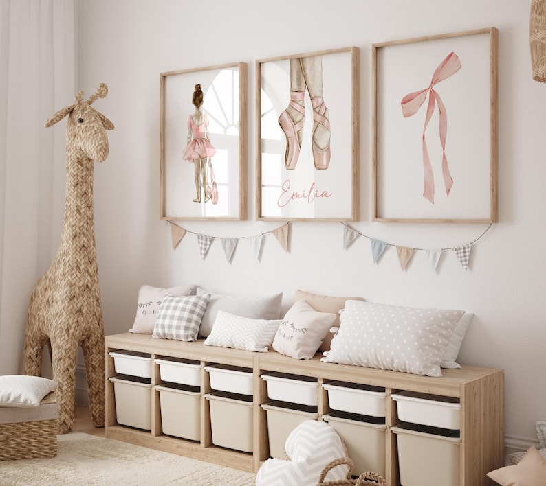 May include: A light wood bench with white storage bins and three pillows. The bench is decorated with a garland of flags and three framed prints. The prints feature a ballerina, a pair of pink ballet shoes, and a pink ribbon.