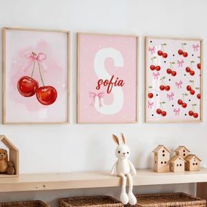 Puede incluir: Tres impresiones enmarcadas en rosa y blanco con un tema de cereza. La primera impresión presenta dos cerezas rojas con tallos verdes y lazos rosas sobre un fondo rosa con lunares blancos. La segunda impresión presenta una letra S blanca con el nombre "sofia" en letras rojas sobre un fondo rosa con lunares blancos. La tercera impresión presenta un patrón repetitivo de cerezas rojas con tallos verdes y lazos rosas sobre un fondo rosa con lunares blancos y corazones negros.
