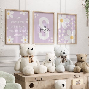 May include: Three framed prints with a purple background and white daisy flowers. The center print has a large white letter "S" with the name "Sofia" written in black cursive. The prints are hanging on a white wall above a wooden shelf with four teddy bears.